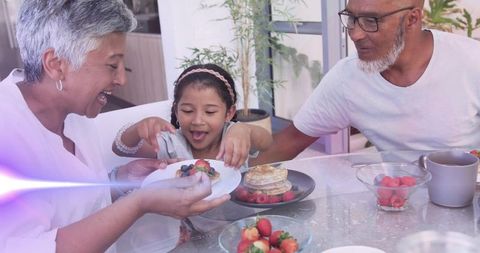 Grandparents sharing joyful breakfast with granddaughter offering berries and pancakes