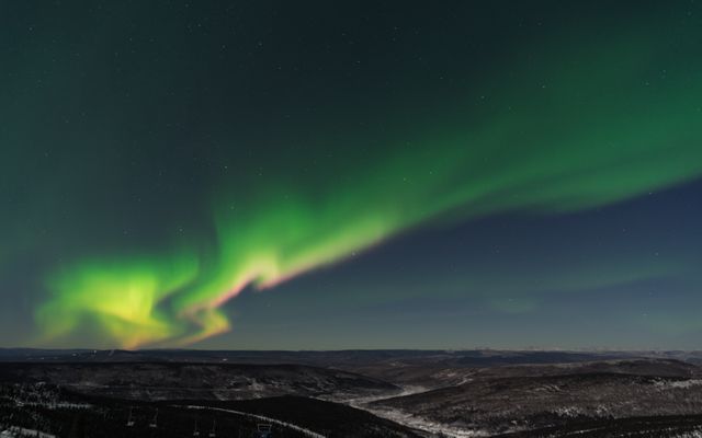 Northern lights illuminating dark sky with green and yellow hues over snowy mountainous region. Perfect for travel blogs, nature documentaries, environmental awareness campaigns, and atmospheric scenic backgrounds.