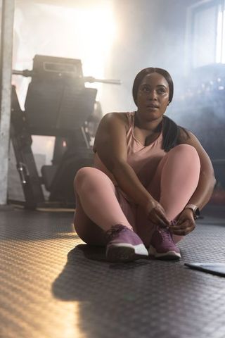 African american woman tying shoelaces in gym wearing pink activewear and smartwatch, backlit studio