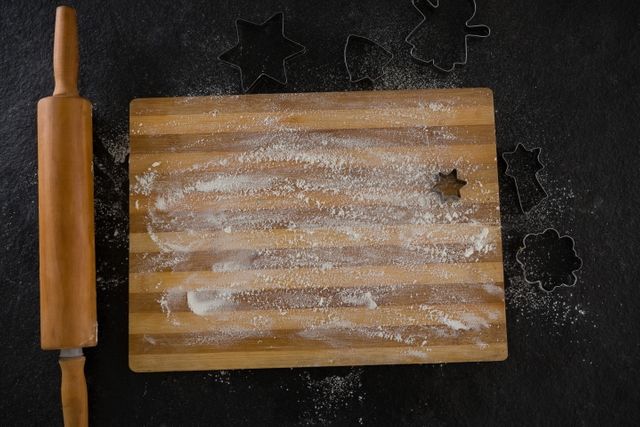 Flour-dusted chopping board with various cookie cutters and a rolling pin on a black background. Ideal for illustrating baking, cooking preparation, and kitchen activities. Suitable for use in food blogs, recipe books, and culinary websites.