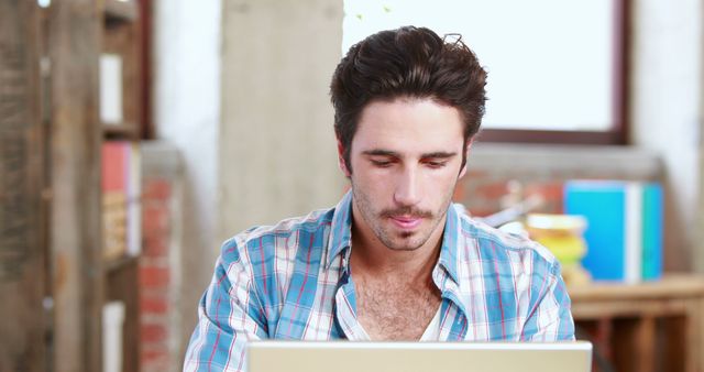 Young man concentrating while working on a laptop. Perfect for illustrating modern technology use, remote work, young professional lifestyle, educational content, office environments, freelance work topics, and indoor working spaces.