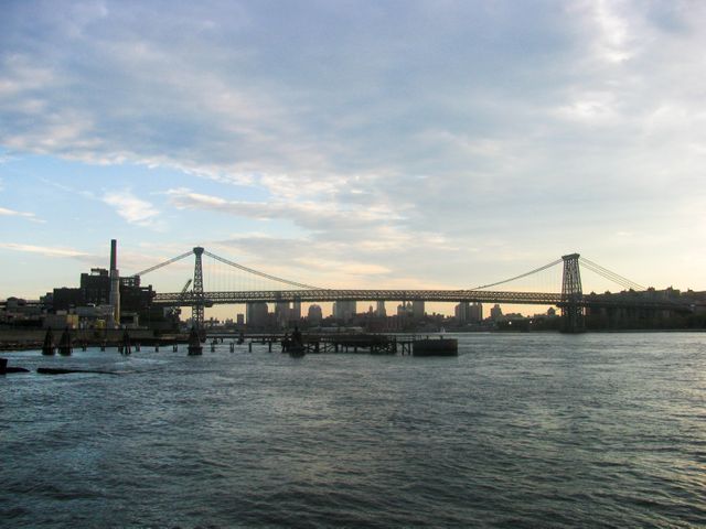 This iconic city scene features a suspension bridge spanning a river, with industrial buildings and a skyline in the background. The sky is partly cloudy, adding texture to the serene late afternoon setting. Ideal for projects involving urban life, infrastructure, travel promotions, postcards, and architectural studies. Additionally, the soft lighting lends a calm ambiance that suits inspirational content and city-themed art.