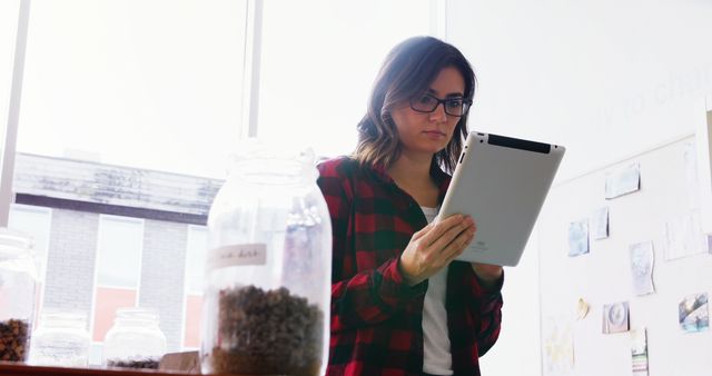 Young woman in casual attire with glasses using tablet to check inventory in office with natural light. Large glass jars filled with items on the desk. Ideal for use in business, technology, and productivity contexts, or to illustrate modern workspace habits.