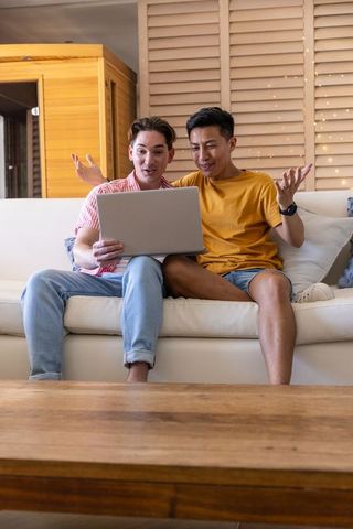 Multiracial friends sharing laptop on sofa, laughing and reacting in cozy living room
