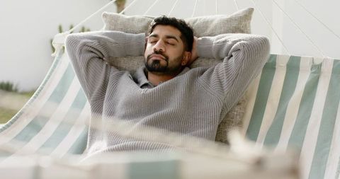 Indian man reclining on striped hammock relaxing outdoors with cushion and cozy sweater
