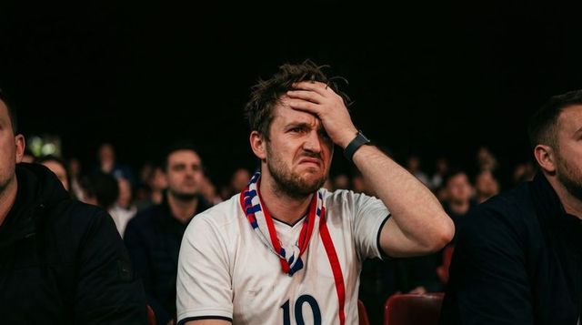 Despairing fan holding head while watching tense football match in crowded stadium with scarf
