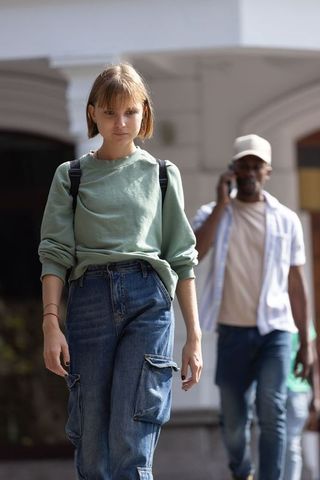 Young woman walking through sunlit urban archway with backpack while man talks on phone