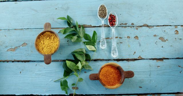 Various spices including turmeric and red peppercorns are arranged on a rustic blue wooden table, with copy space. The vibrant colors of the spices add a warm and inviting aesthetic to the image.