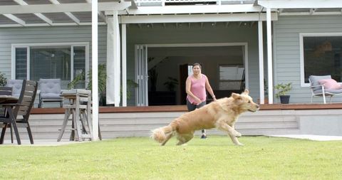 Mature woman watching golden retriever running across backyard lawn near contemporary patio