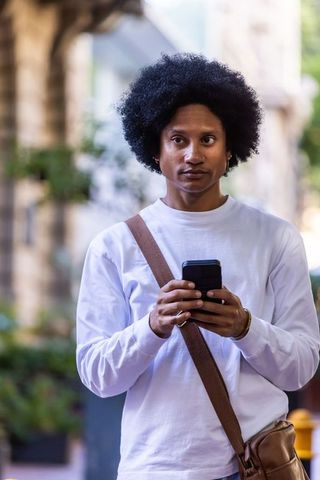 African american man holding smartphone on city sidewalk wearing white shirt and satchel