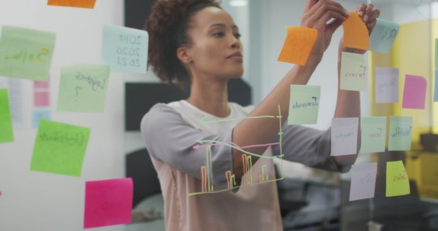Young businesswoman organizing colorful sticky notes on transparent board in modern office. Useful for content on project management, teamwork, business planning, innovation, and corporate strategy.