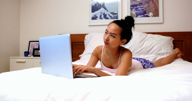 Young woman in casual attire looks focused while browsing on a laptop in a cozy bedroom. This could be used for themes related to technology, remote work, e-learning, home relaxation, and digital lifestyles.