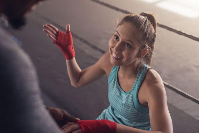 Female boxer smiling while coach wraps her hands in gym. Ideal for use in fitness and sports-related content, promoting teamwork, strength training, and athletic preparation. Suitable for articles, advertisements, and social media posts about boxing, personal training, and gym environments.