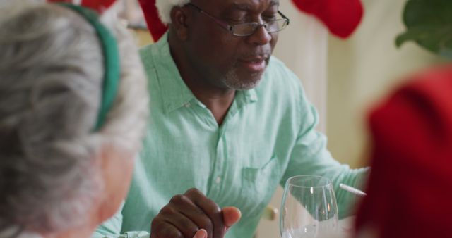 Happy african american senior man praying with friends, celebrating meal at christmas time. christmas festivities, celebrating at home with friends.