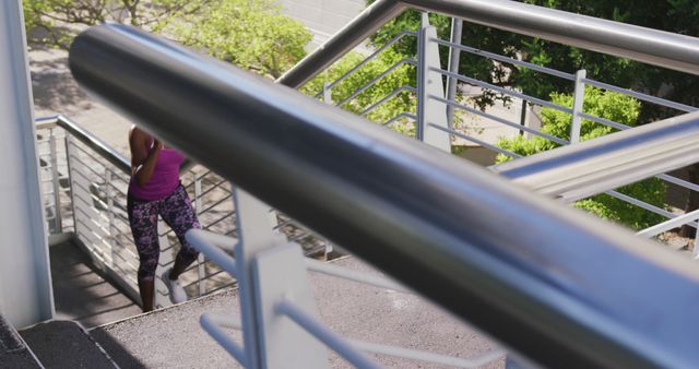 Woman seen climbing an outdoor staircase wearing workout clothes. Sunlight filters through trees creating a vibrant atmosphere ideal for visual content related to fitness, exercise, urban lifestyles, and active living. Perfect for websites, fitness blogs, and advertising campaigns promoting staying fit in urban environments.