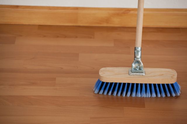 This image shows a close-up of a broom with blue bristles and a wooden handle sweeping a wooden floor. Ideal for use in articles or advertisements related to household cleaning, maintenance, and hygiene. Suitable for illustrating concepts of cleanliness and domestic chores.