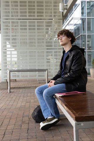 Teenage student sitting on bench at modern campus plaza with backpack and notebooks