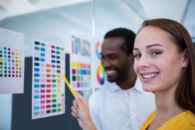 This image shows two graphic designers collaborating over color charts in a modern office. The female designer is smiling and pointing at the chart, while the male designer is engaged in the discussion. This image can be used to depict teamwork, creativity, and the design process in a professional setting. Ideal for articles, blogs, or websites related to graphic design, creative industries, and office collaboration.