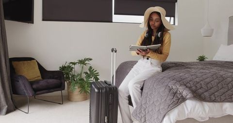 Young woman preparing for trip in modern hotel, sitting on bed with suitcase and tablet