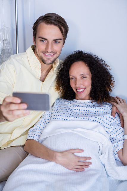 Expectant couple capturing a joyful moment in a hospital ward. The woman is in a hospital gown, indicating she is likely in labor or preparing for childbirth. This image can be used for themes related to pregnancy, maternity care, hospital experiences, family bonding, and modern technology in healthcare settings.
