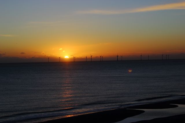 Depicts a peaceful sunset over an ocean with a row of wind turbines on the horizon, blending natural beauty with renewable energy technology. Great for topics on environmental awareness, renewable energy, sustainable living, peaceful scenes, nature beauty, and technological advancements.