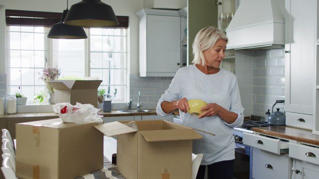 Senior caucasian woman is unpacking boxes in her new kitchen, creating a sense of excitement and change. She is carefully removing crockery from a cardboard box suggesting the start of her new domestic life. Ideal for themes related to relocation, retirement, homemaking tips, real estate advertising, or home organization and lifestyle changes.