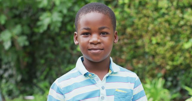 This image features a young boy standing in an outdoor setting, dressed casually in a striped shirt with a subtle smile on his face. The lush greenery in the background adds a natural and vibrant element to the scene. Ideal for uses in educational materials, children's fashion ads, lifestyle blogs, and family-centered campaigns.