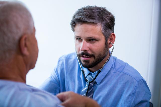 A male doctor in a blue shirt and tie is attentively listening to a senior patient's heartbeat with a stethoscope in a hospital or clinic. Ideal for illustrating medical examinations, healthcare services, doctor-patient relationships, medical consultations, and professional healthcare environments.