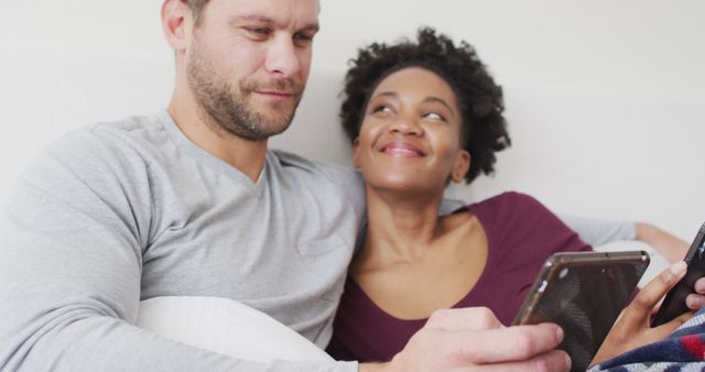 Multicultural couple relaxing together at home, each using their smartphones. Woman smiling warmly, enjoying time with her partner. Scene captures everyday life, tech use, and happiness in a relaxed, cozy setting. Suitable for lifestyle, technology, relationships, and home living concepts.