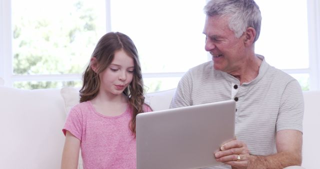 Grandfather and granddaughter are sitting on a white couch using a laptop together. The elderly man, wearing a gray shirt, smiles while the girl, in a pink shirt, concentrates on the screen. Suitable for themes related to family bonding, technology usage across generations, home environment, and elderly teaching youth.