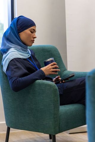 Young professional woman in blue hijab relaxing with tumbler and tablet in modern lounge