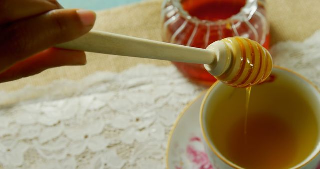 Sweet honey is seen drizzling from a wooden honey dipper into a cup of tea. In the background, a jar of honey sits on a table covered with a lace tablecloth. The image showcases the use of honey as a natural sweetener in hot beverages. This could be used in culinary magazines, health-and-wellness blogs, morning routine visuals, and recipes involving honey in drinks.