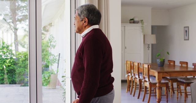 Elderly man gazing out a large window in a cozy home interior, dressed in a maroon sweater. Well-lit dining area with wooden table and chairs in the background adding to the cozy atmosphere. This image can be used to represent themes of retirement, contemplation, or nostalgia in various contexts such as dedicated blogs, elder care articles, or lifestyle websites.