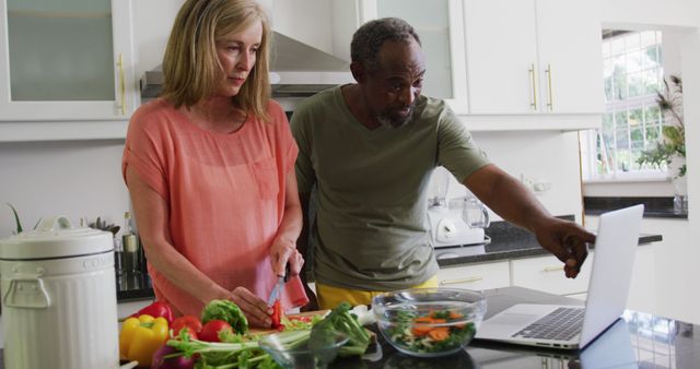 Mature couple preparing vegetables in a modern kitchen while referring to a laptop for recipes or instructions. Ideal for articles and content related to healthy living, collaborative cooking, and staying updated with technology in everyday tasks.