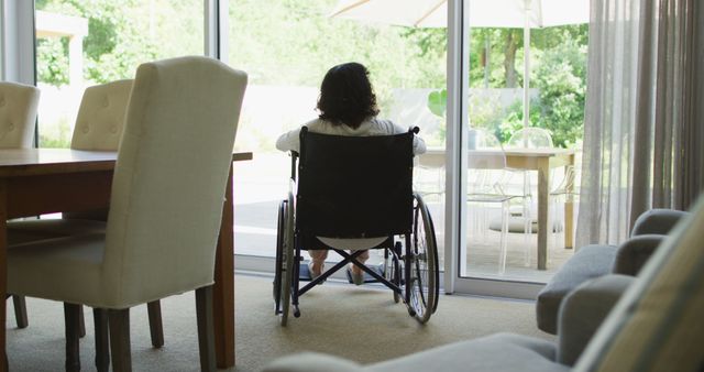Biracial disabled woman in wheelchair drinking coffee and looking out of window in sunny living room. wellbeing and domestic lifestyle with physical disability.