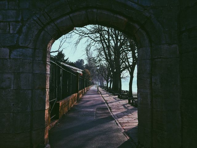 Image features a historic stone archway opening onto a scenic walkway lined with trees. This photo can be used for travel blogs, urban exploration websites, history articles, or architectural highlights.
