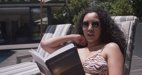 African american woman reading poolside in zebra bikini relaxing on sunlit deck lounger