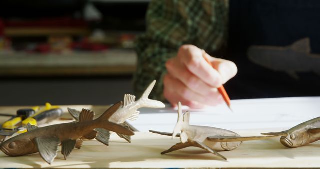 A middle-aged person is sketching on paper, surrounded by wooden fish sculptures, with copy space. Their focus on the drawing suggests a creative or artistic profession, a sculptor or a designer planning their next piece.
