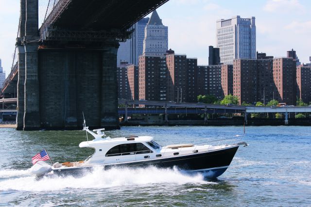 Luxury yacht cruising along river beneath large bridge in bustling urban area. Tall skyscrapers line waterfront in sunny, clear day. Ideal for travel blogs, advertisement for boating services, or urban adventure narratives.