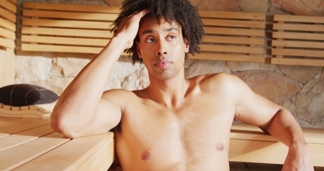 Image of relaxed bare chested biracial man sitting in sauna room at health spa. Vacation, relaxation, health and inclusivity concept.