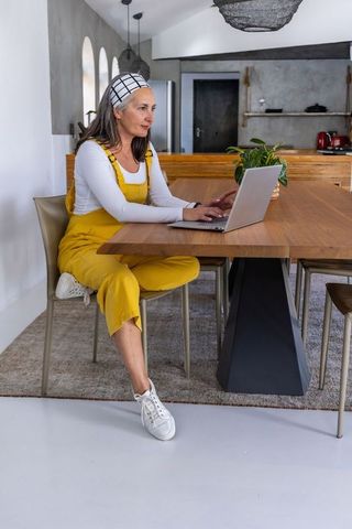 Middle-aged woman working on laptop at dining table in modern open-plan kitchen