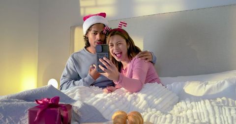 Asian couple wearing festive hats taking cozy holiday bed selfie with gift