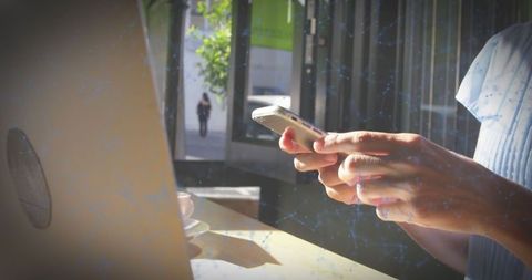 Female Working Remotely Using Smartphone and Laptop at Sunlit Cafe Table During Coffee Break
