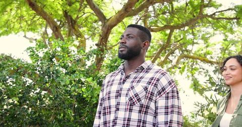 Couple enjoying sunlit park with man gazing upward under lush tree canopy, woman smiling