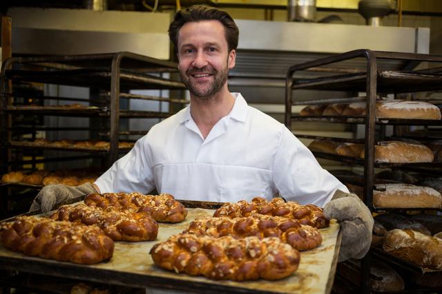 Smiling baker holding tray of freshly baked twisted buns in a bakery kitchen. Ideal for use in culinary blogs, bakery advertisements, cooking websites, and food-related marketing materials.