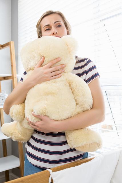 Woman standing indoors holding large teddy bear, looking sad and contemplative. Natural light from window illuminates scene, creating a somber mood. Useful for topics on mental health, grief, motherhood, emotional support, and coping with loss.
