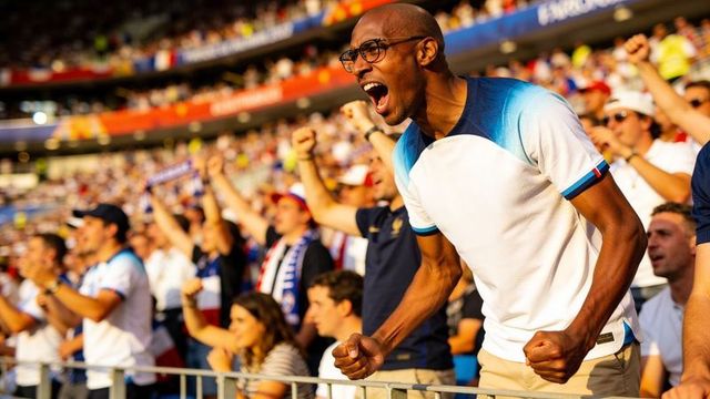 Cheering male fan wearing glasses and white blue shirt leaning on railing in stadium crowd