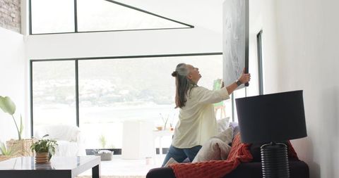 Mature woman hanging large framed art in modern waterfront living room with natural light