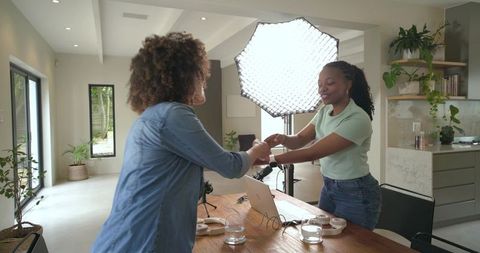 Female content creators shaking hands in bright home studio with laptop and softbox