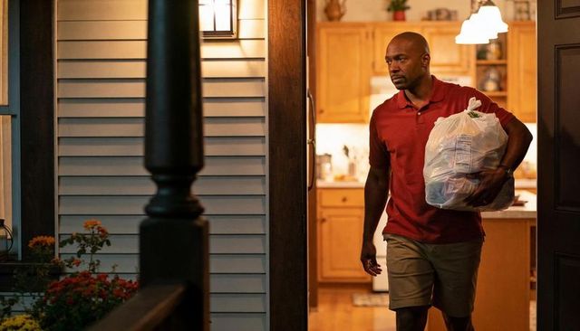 Man carrying grocery bag through front doorway into warmly lit kitchen at dusk